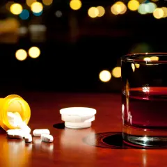 Nalany ibuprofen i szklanka alkoholu na drewnianym blacie, w tle rozmyte światła.
