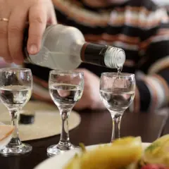 Ręka nalewa wódkę do kieliszków, tworząc potencjalny rekord promili. Na stole wędlina i przekąski.