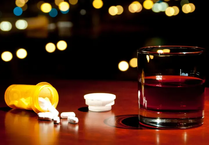 Nalany ibuprofen i szklanka alkoholu na drewnianym blacie, w tle rozmyte światła.