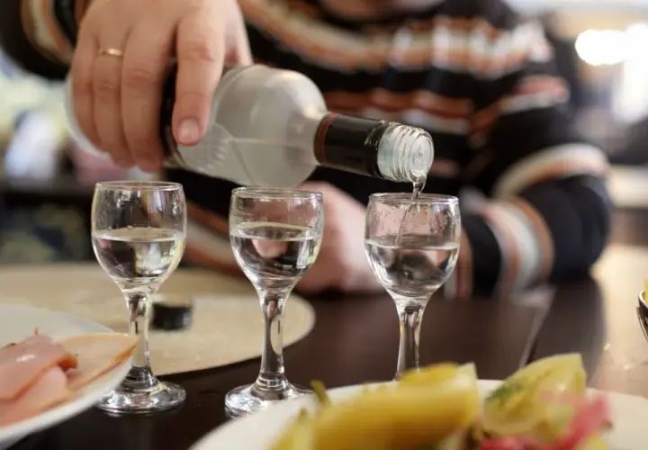 Ręka nalewa wódkę do kieliszków, tworząc potencjalny rekord promili. Na stole wędlina i przekąski.