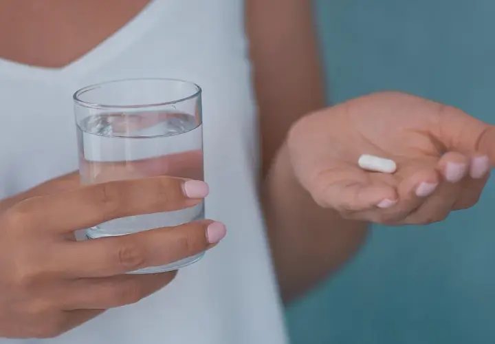 Dłoń trzyma szklankę wody i białą kapsułkę, która może być jednym z neuroleptyków.