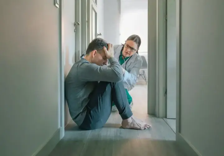 Lekarz wspiera pacjenta zmagającego się ze splątaniem w psychiatrii. Mężczyzna siedzi na podłodze, przygnębiony, a lekarka pochyla się nad nim z troską.