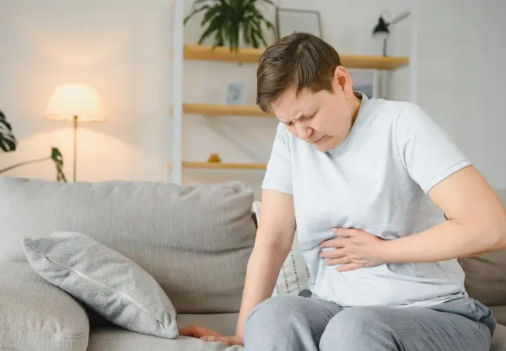 Kobieta siedzi na kanapie, trzymając się za brzuch. Jej twarz wyraża ból, co może sugerować przewlekłe zapalenie trzustki.
