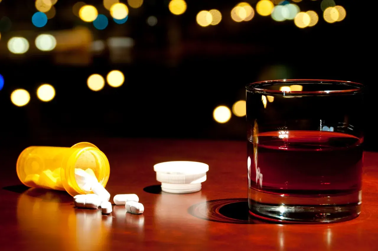 Nalany ibuprofen i szklanka alkoholu na drewnianym blacie, w tle rozmyte światła.