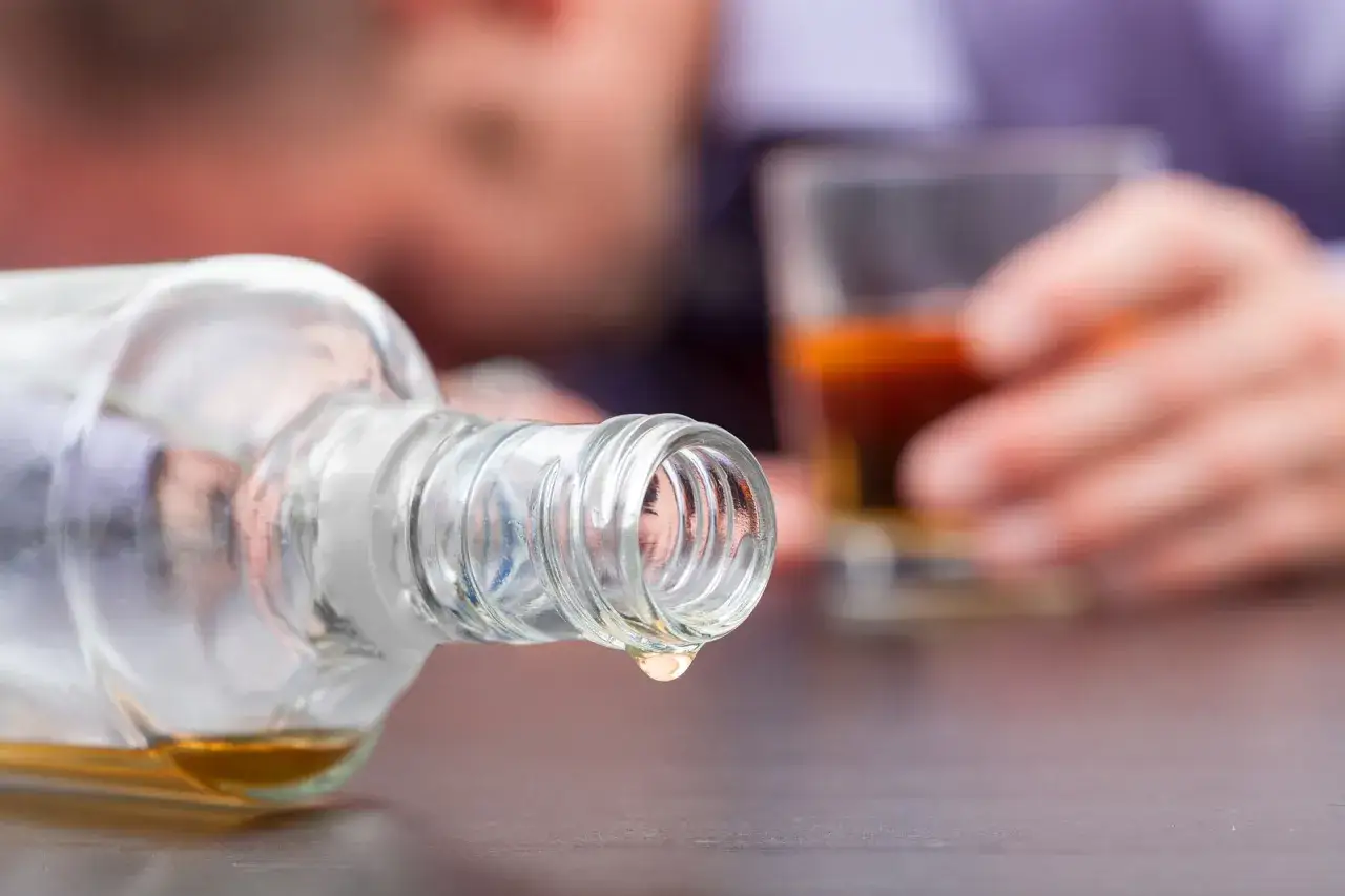 Kropla alkoholu kapie z butelki. W tle widać osobę z kieliszkiem, być może pijącą metanol.