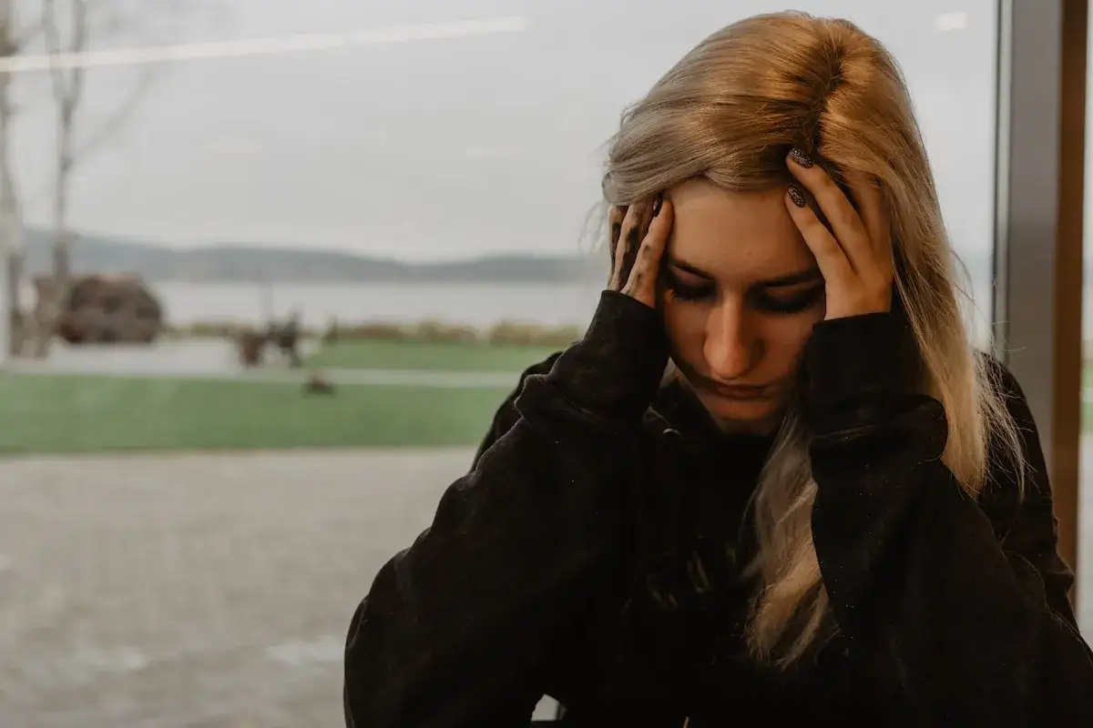 Młoda kobieta z blond włosami trzyma się za głowę, jej twarz wyraża cierpienie. Może to być objaw zespołu serotoninowego.